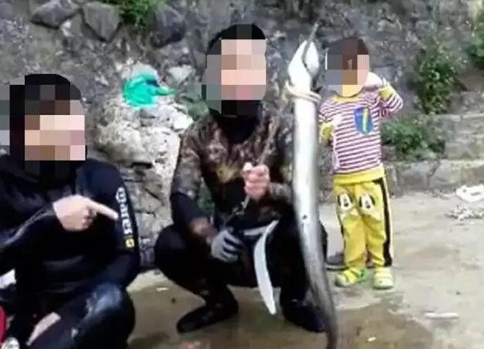 一名穿着迷彩潜水服的男子脸上被打上了马赛克,他手里握着一根金属棒,而棒的一头则挂着一条鱼,整个神态看起来像是在炫耀“战利品”。